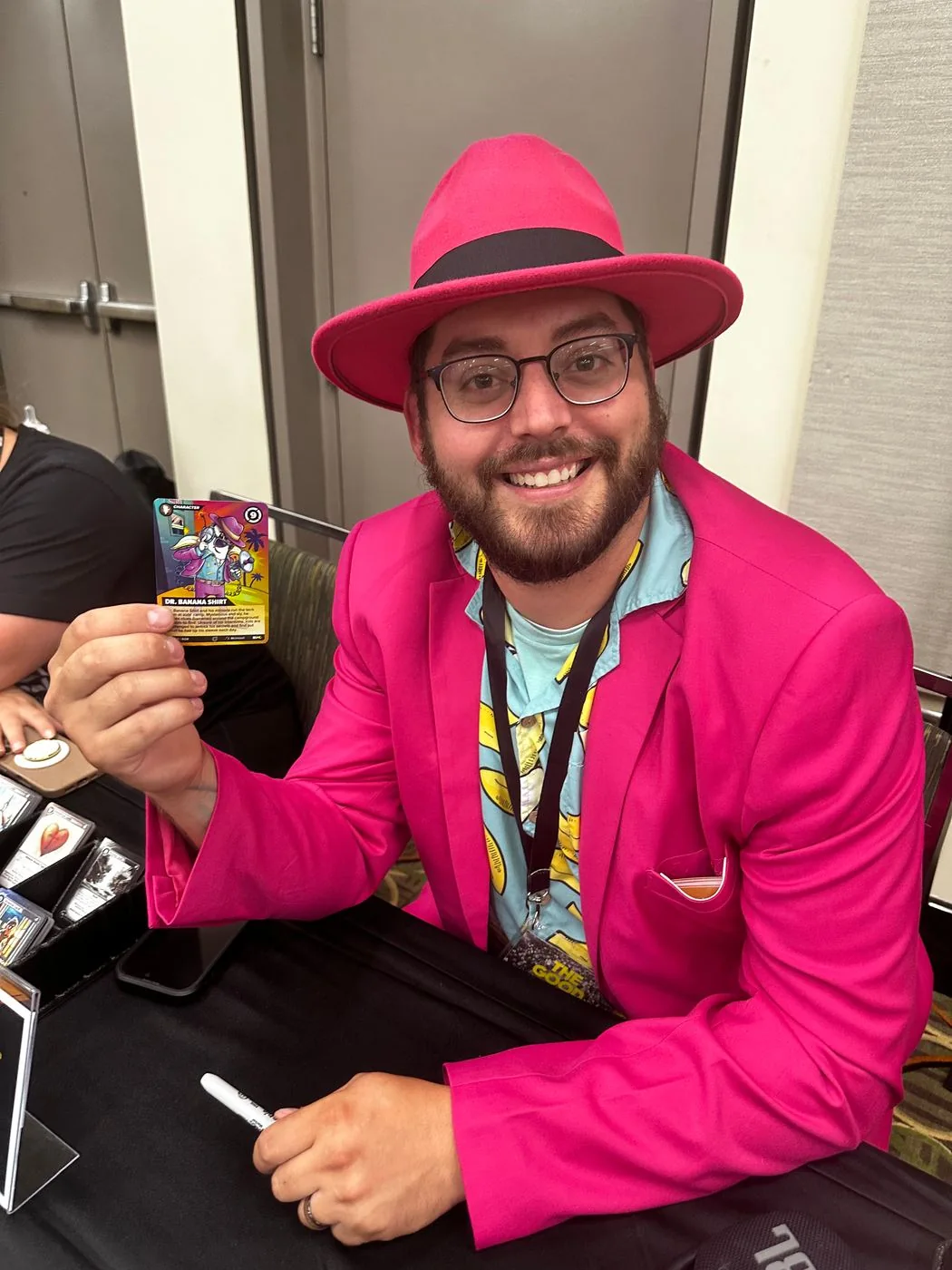 An attendee in a bright pink suit and hat smiles while showing a BGMC character card.
