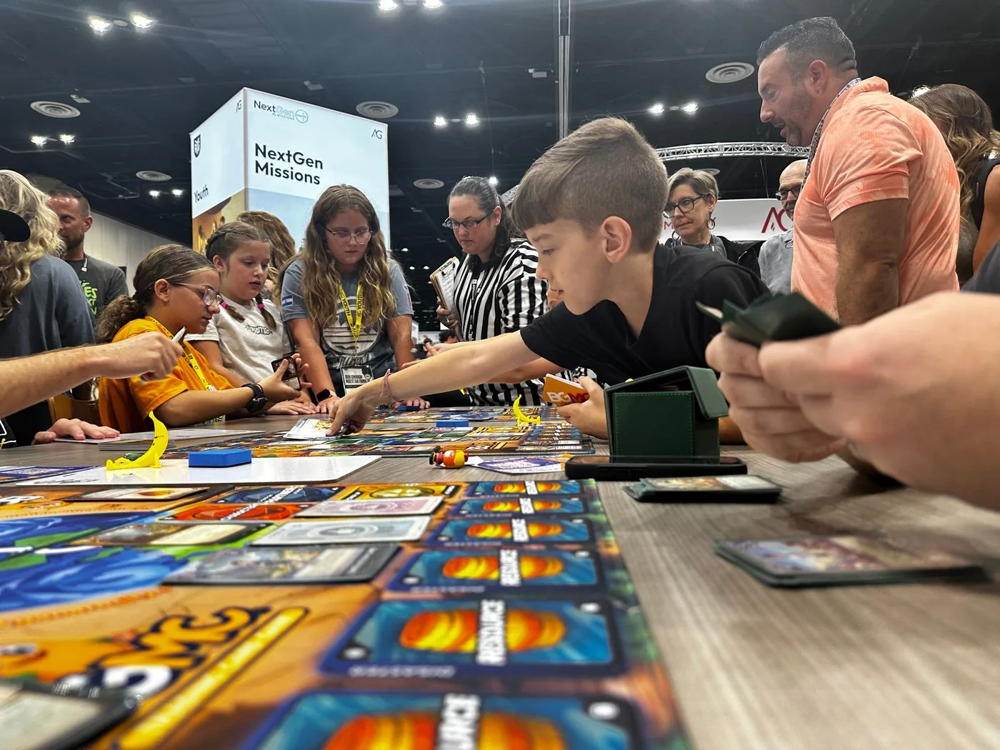 Young players make their next move in a BGMC card game as a referee and families watch.